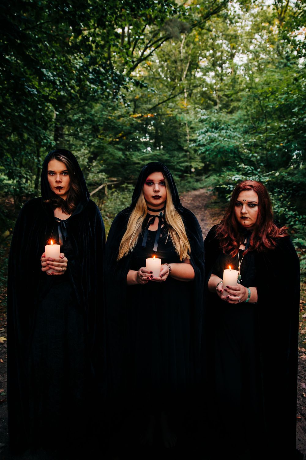 Three figures in black cloaks hold glowing candles in a dark forest setting.