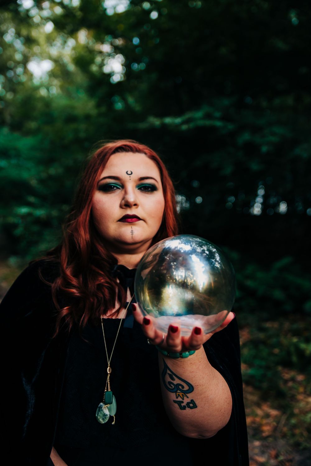 A person in gothic attire holds a crystal ball in a mystical forest setting.