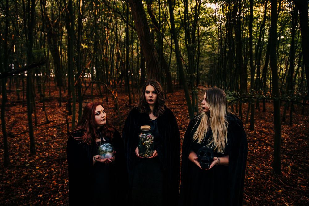 Three figures in black cloaks stand among autumn trees holding mystical orbs.