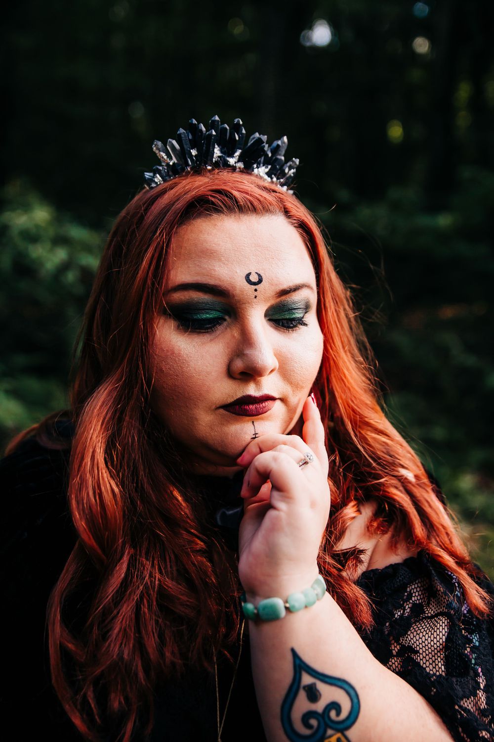 Mystical portrait with black crystal crown and ornate makeup in forest setting.