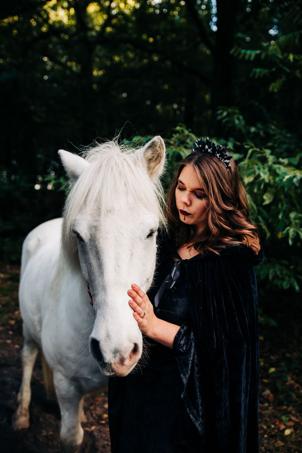 Beautiful white horse and figure in black velvet cloak share a gentle moment in the woods.