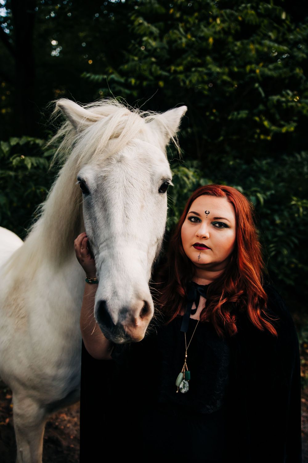 Majestic white horse poses with gothic styled figure in dark forest setting.