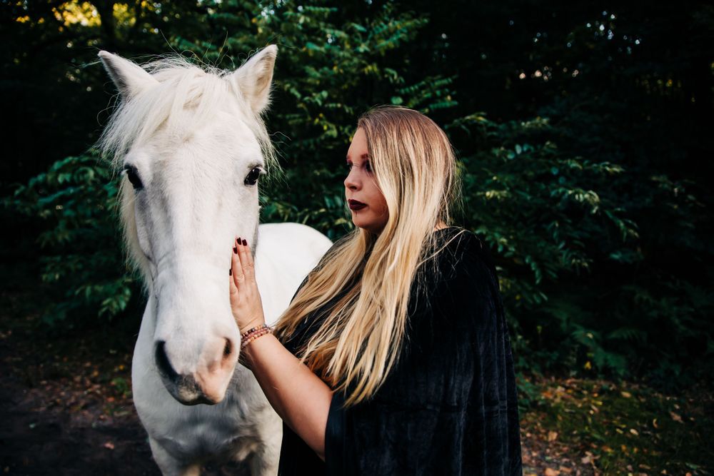 Gentle interaction between white horse and black-cloaked figure in forest.