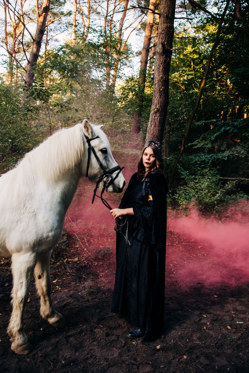 White horse and black-clad figure with pink smoke effects in a moody forest photoshoot.