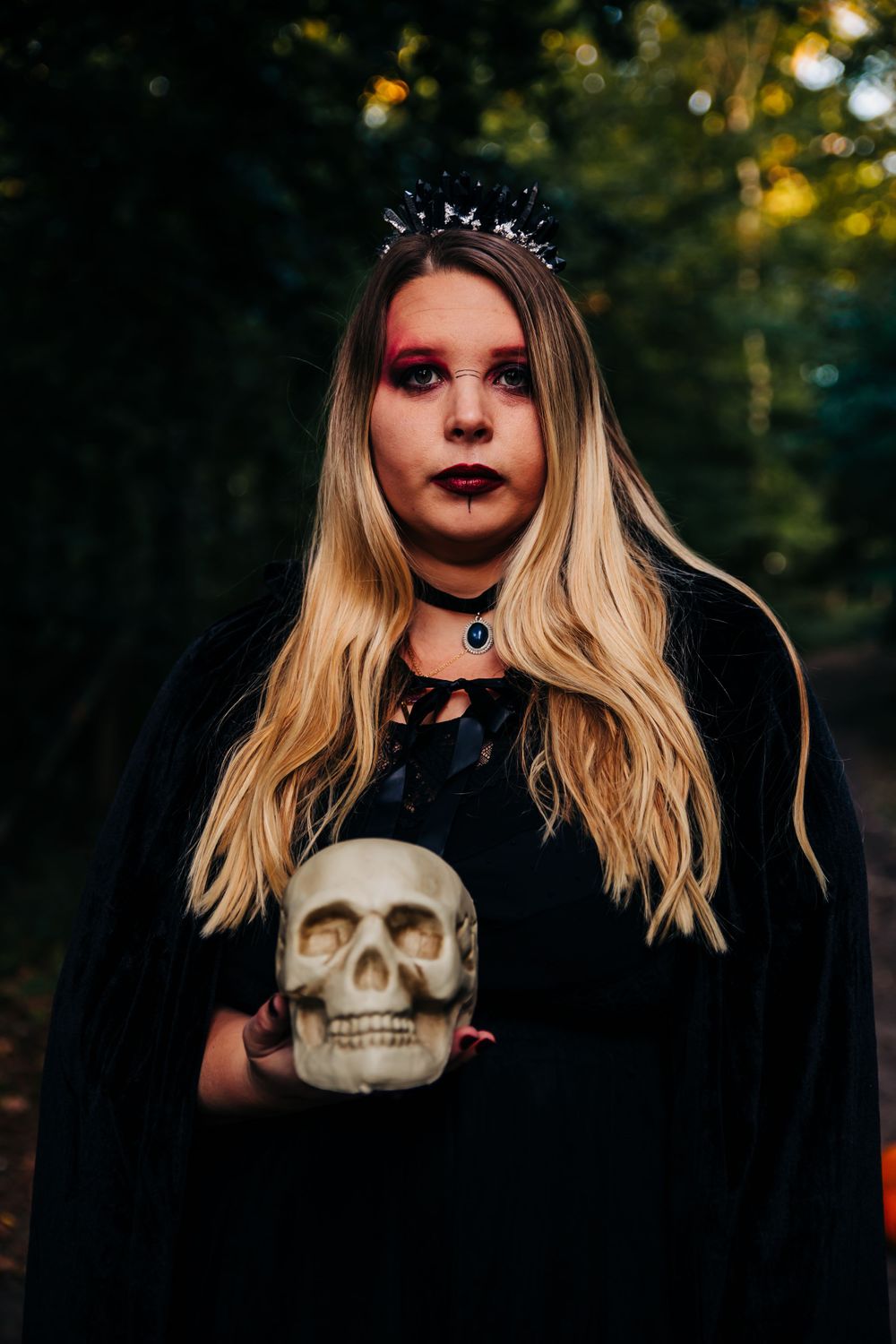 Dark gothic fashion with crystal crown and decorative skull prop in forest setting.
