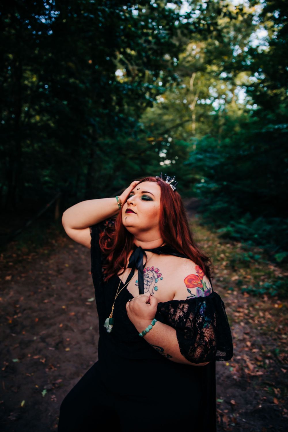 Gothic styled photoshoot in dark forest features black lace dress and visible flower tattoo.