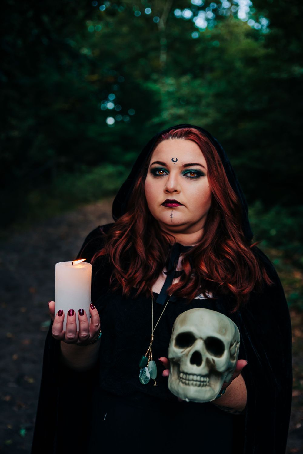 Gothic themed portrait holding skull and candle with dramatic makeup in forest setting.