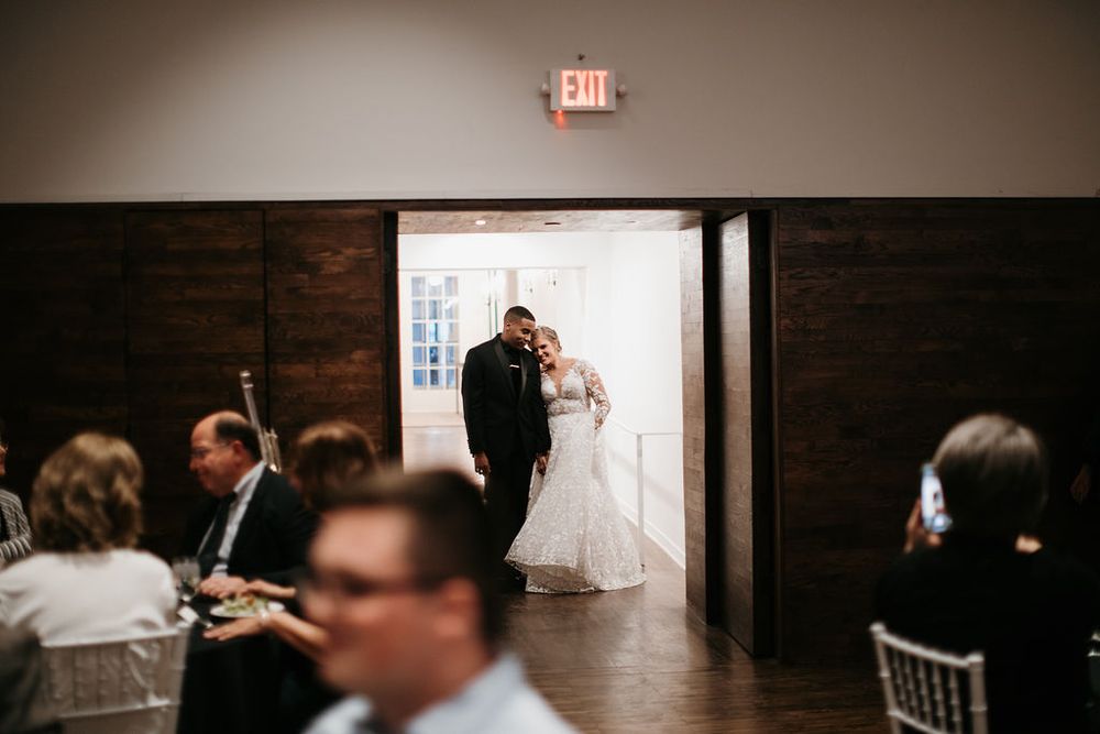 Bride and groom entering wedding reception venue through doorway while guests are seated at tables.