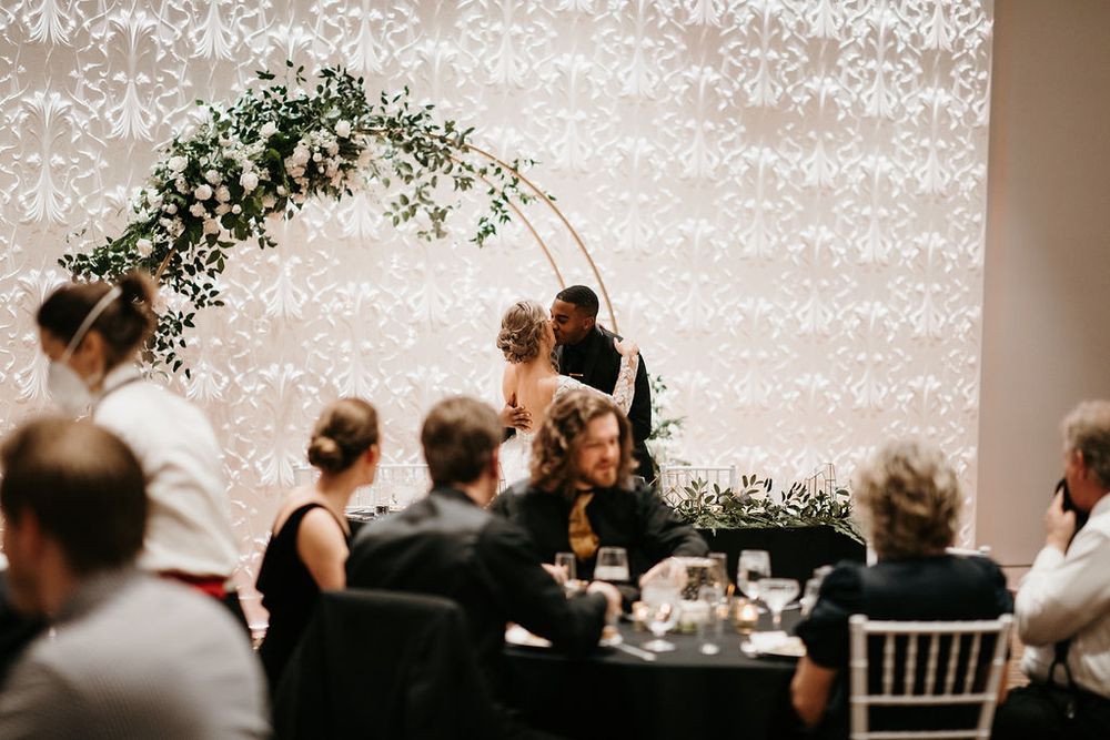 Wedding couple kissing under circular floral arch at sweetheart table during elegant reception.