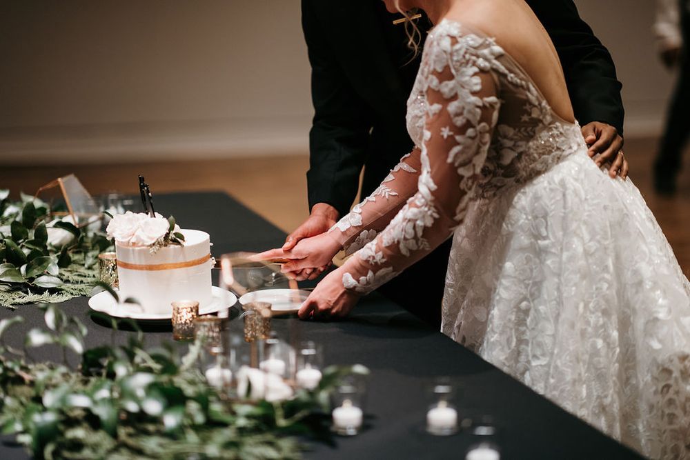 Bride and groom cutting white wedding cake decorated with flowers at reception table with greenery.