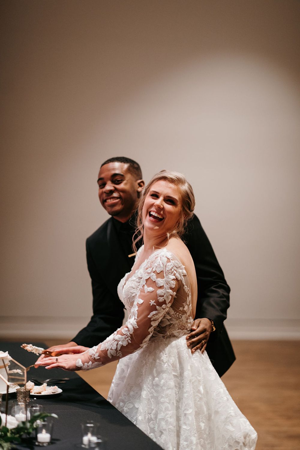 Laughing bride and groom cutting wedding cake together at elegantly decorated reception table.