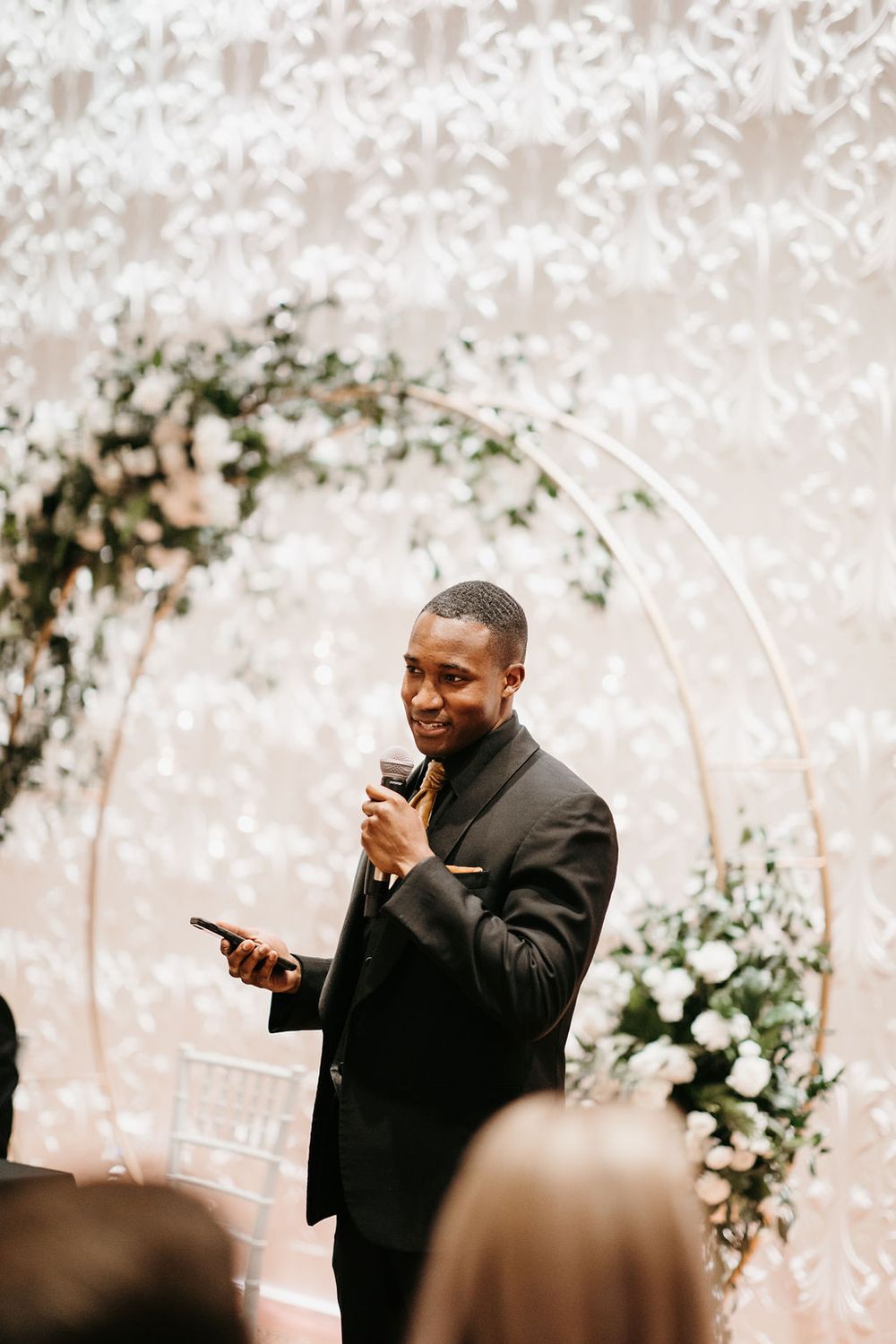 Groomsman delivering speech at wedding ceremony holding microphone near circular floral arch backdrop.