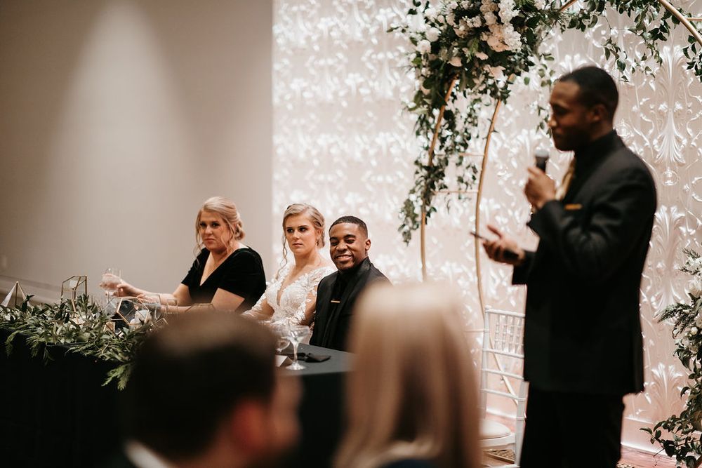 Wedding party seated at sweetheart table listening to guest speech during elegant reception.