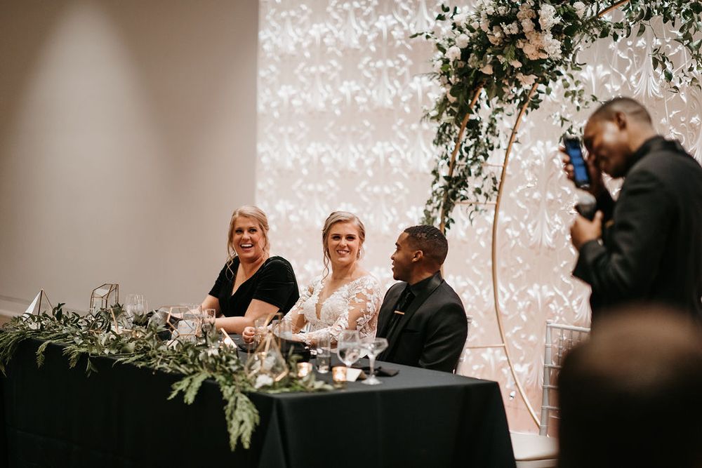 Bride and groom laughing with guest at decorated sweetheart table during wedding reception.