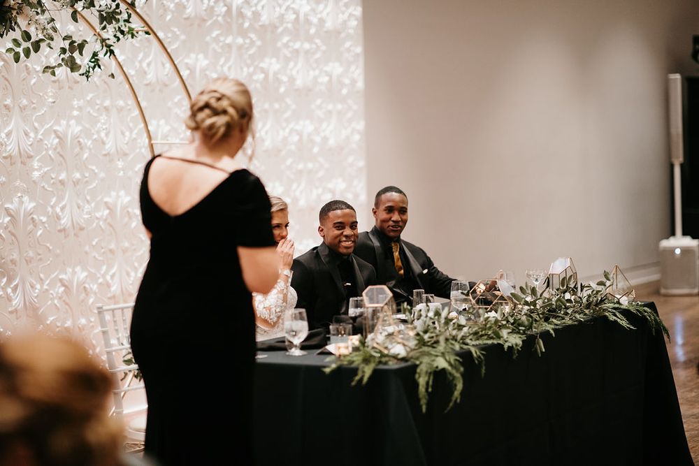 Wedding guests smiling at sweetheart table while woman in black dress delivers reception speech.