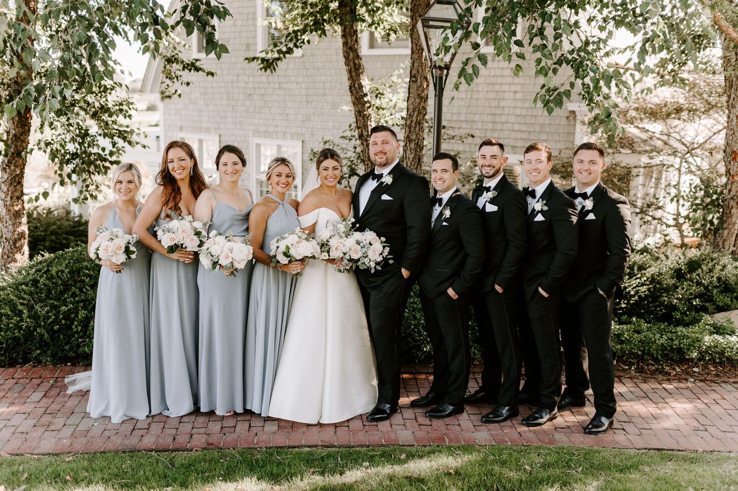 Wedding party poses outdoors with bridesmaids in grey dresses and groomsmen in black tuxedos.
