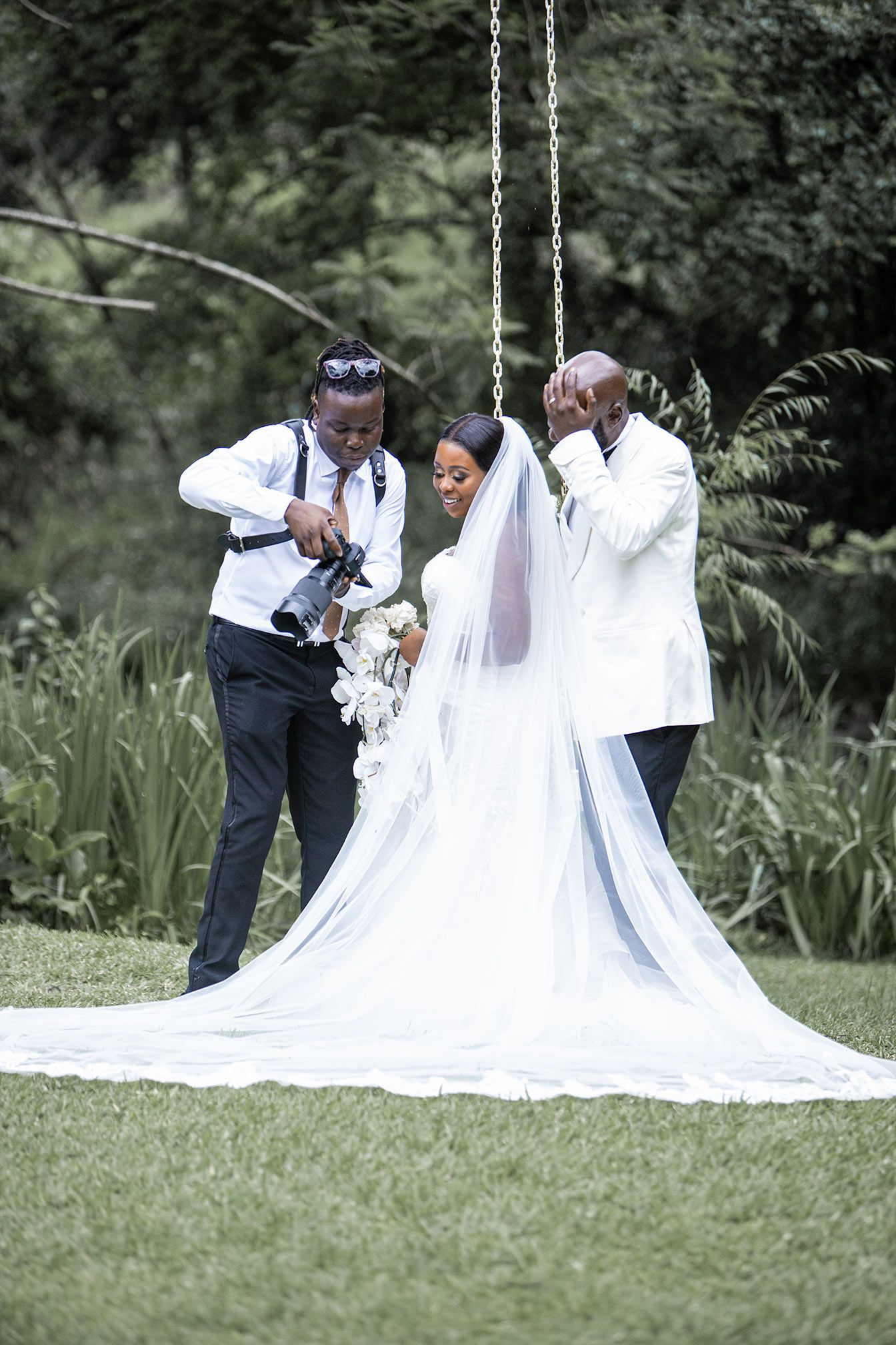 Wedding party helps arrange bride's flowing train outdoors in a garden setting.