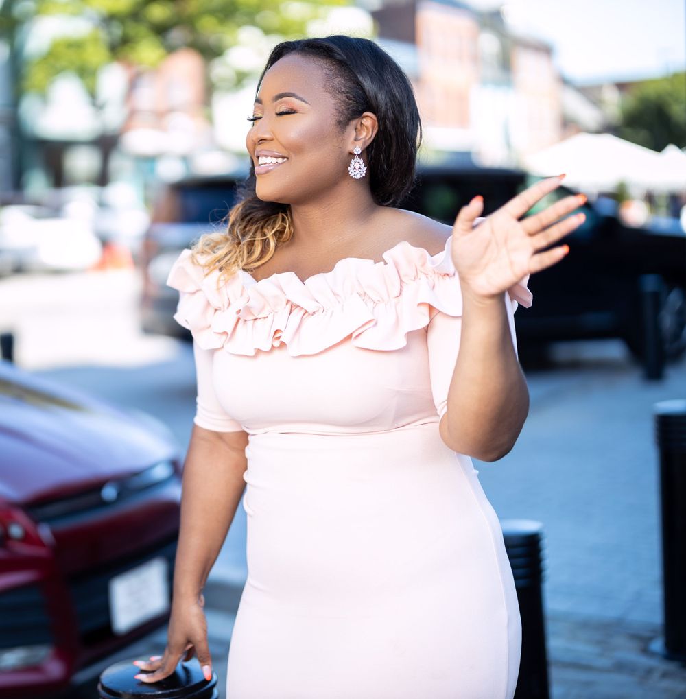 Woman in blush dress waves cheerfully while posing on brick sidewalk.