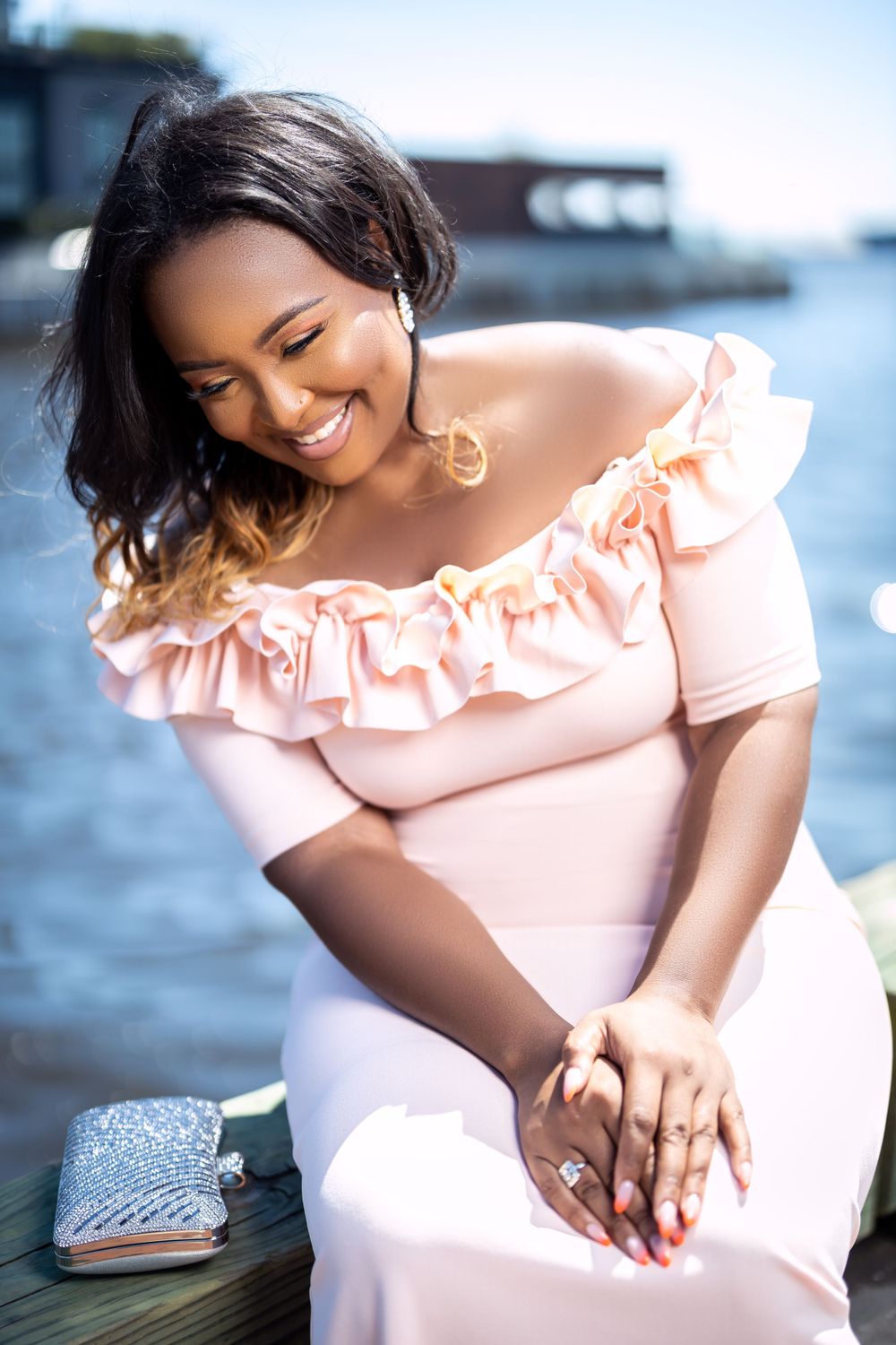 Woman in pink dress sits smiling by waterfront with sparkly purse.