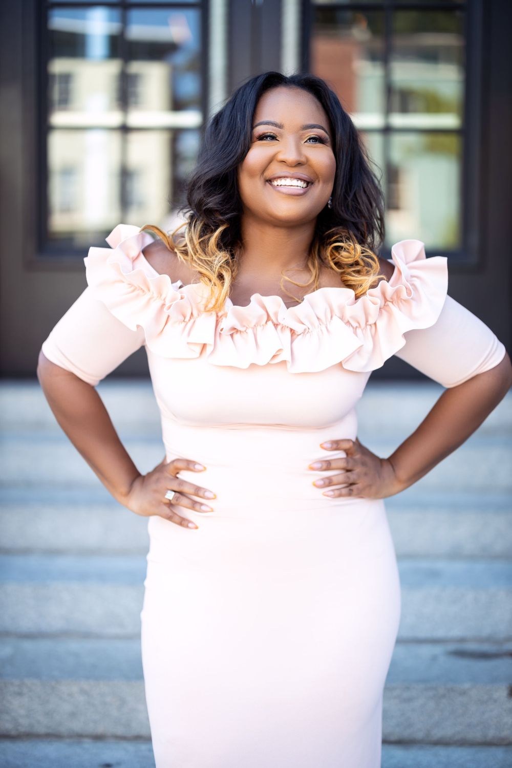 Woman in pink ruffled dress poses confidently with hands on hips outside building.