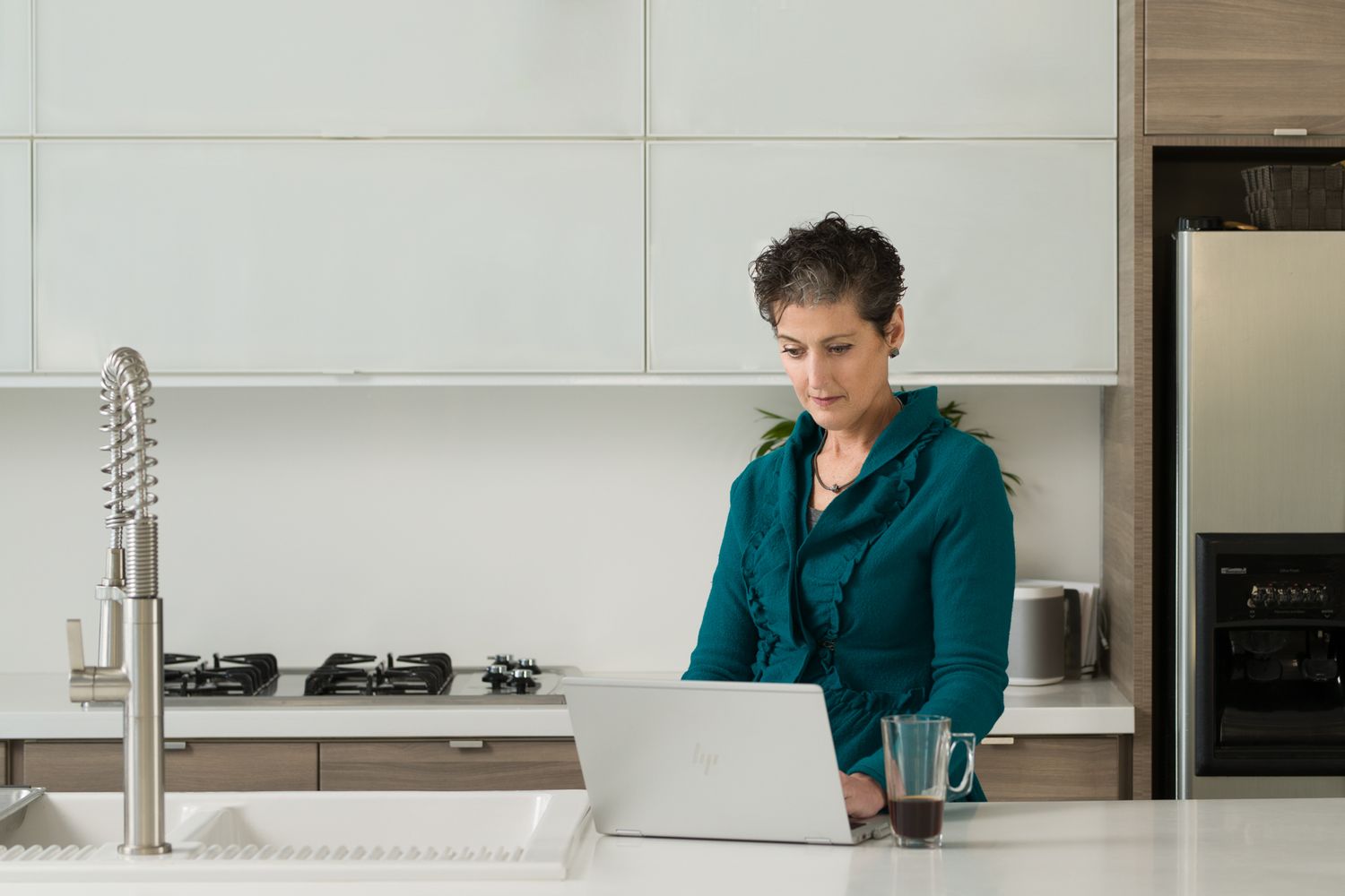Person in teal hoodie working on laptop at modern kitchen counter.