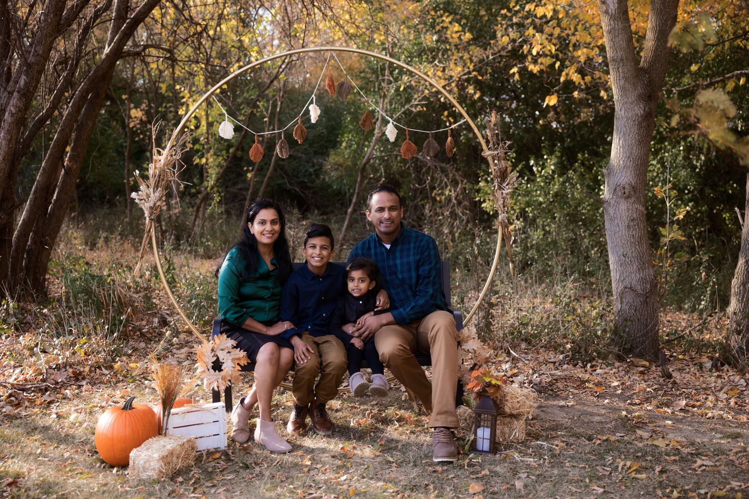 Family sitting with dog under rustic arch with pumpkins and fall decor in outdoor setting.