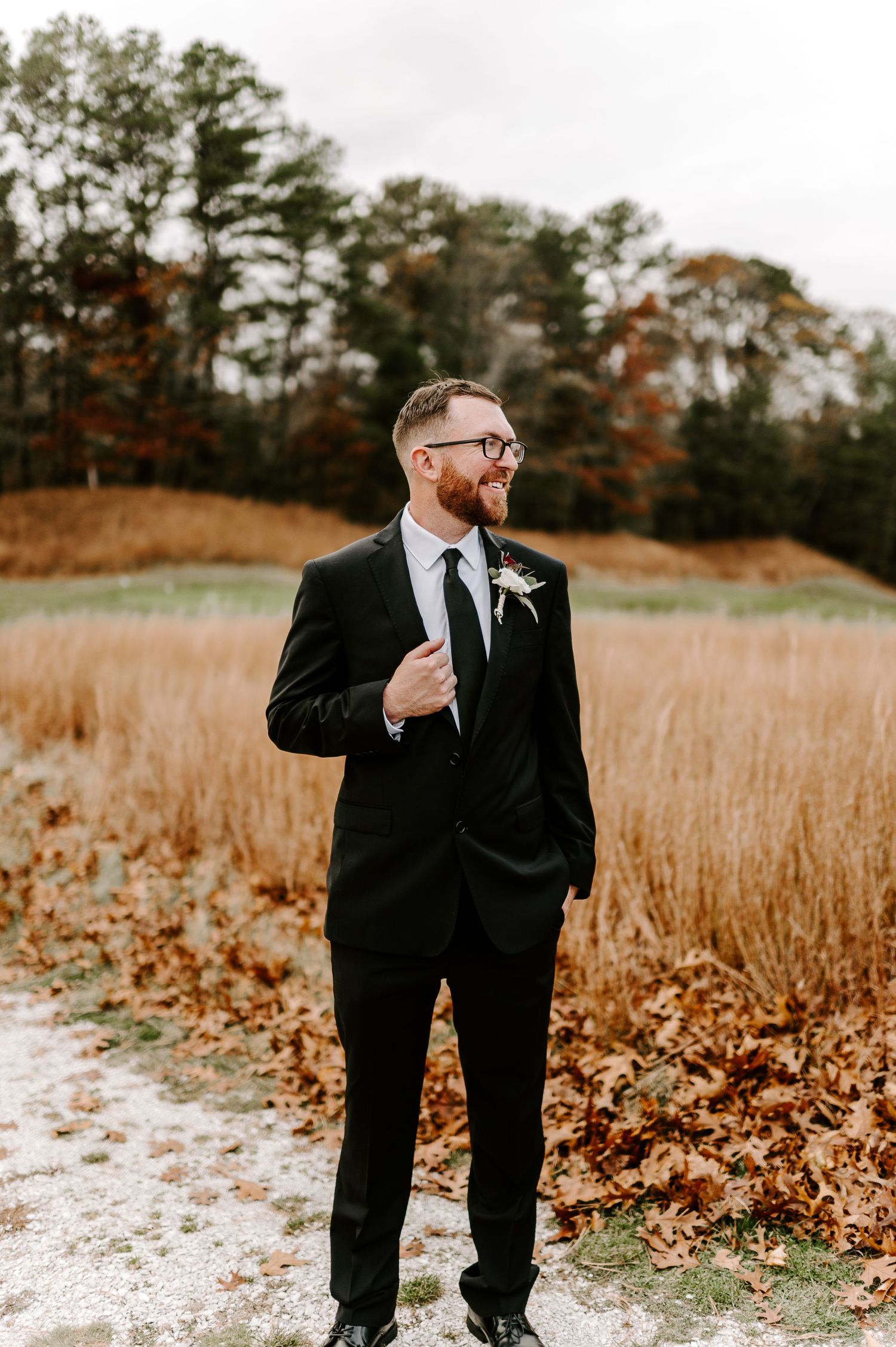 Groomsman in formal attire poses playfully against fall landscape backdrop.