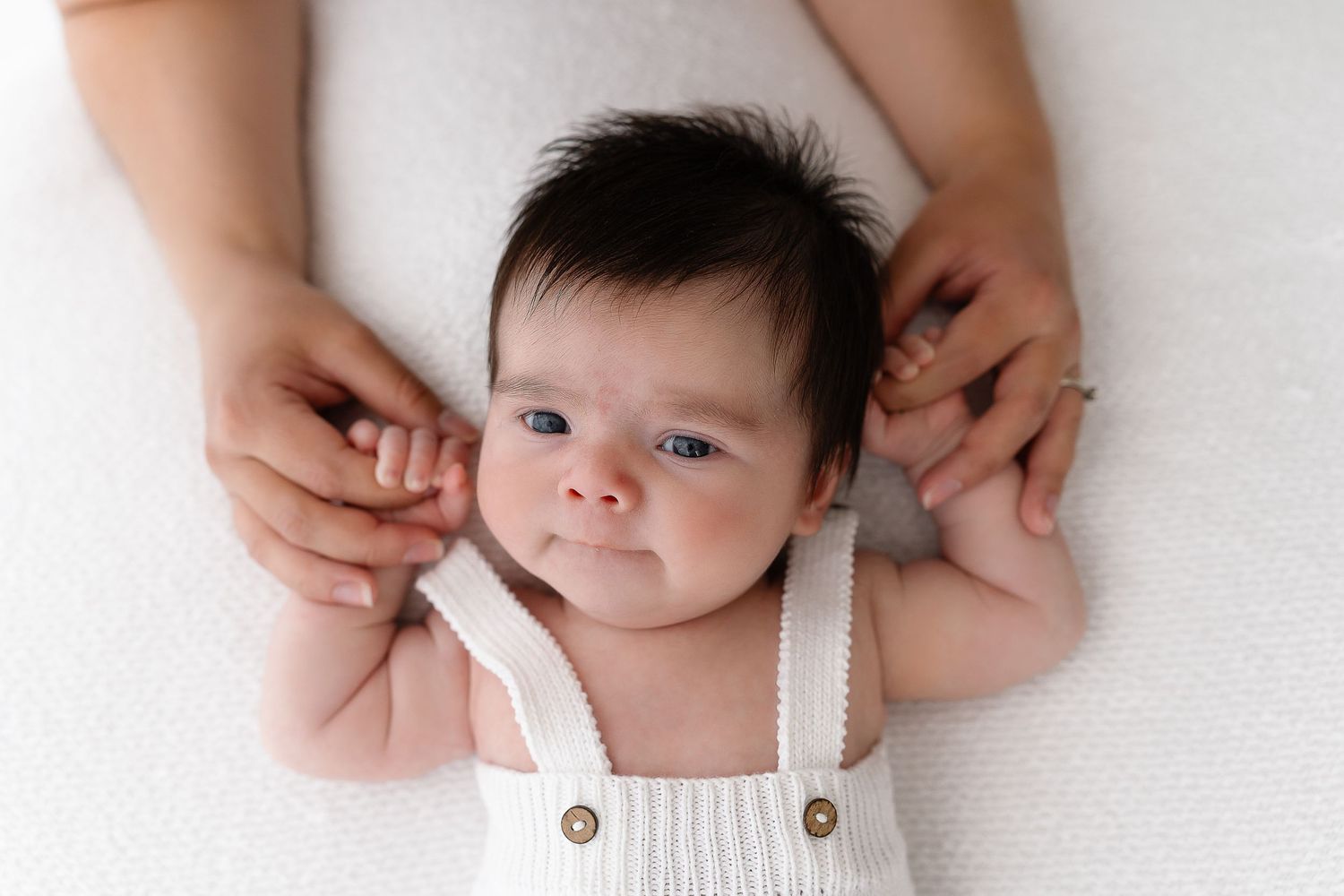 Close-up portrait of baby in white romper with hands framing face.