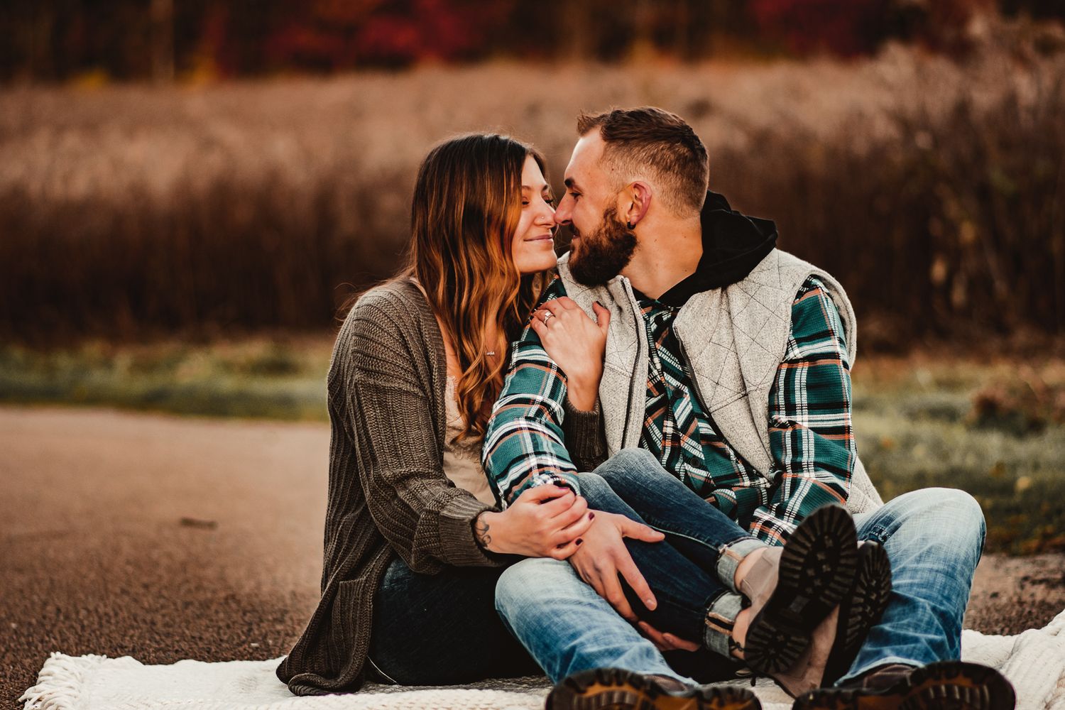 Romantic couple sitting close together on ground wearing plaid and sweaters during golden hour.