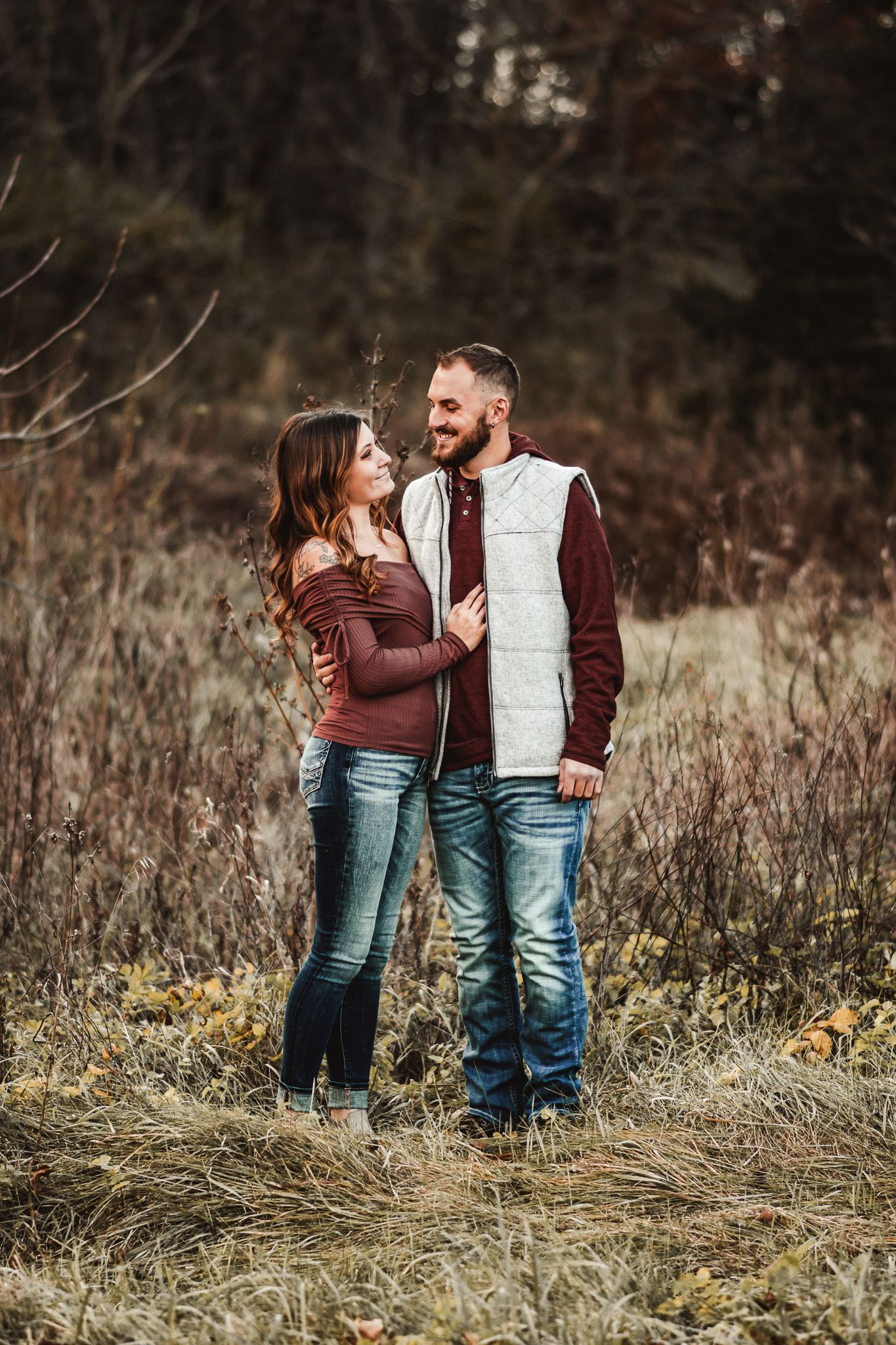A couple in casual fall attire embraces while standing in a field of tall grass during an outdoor photo session.