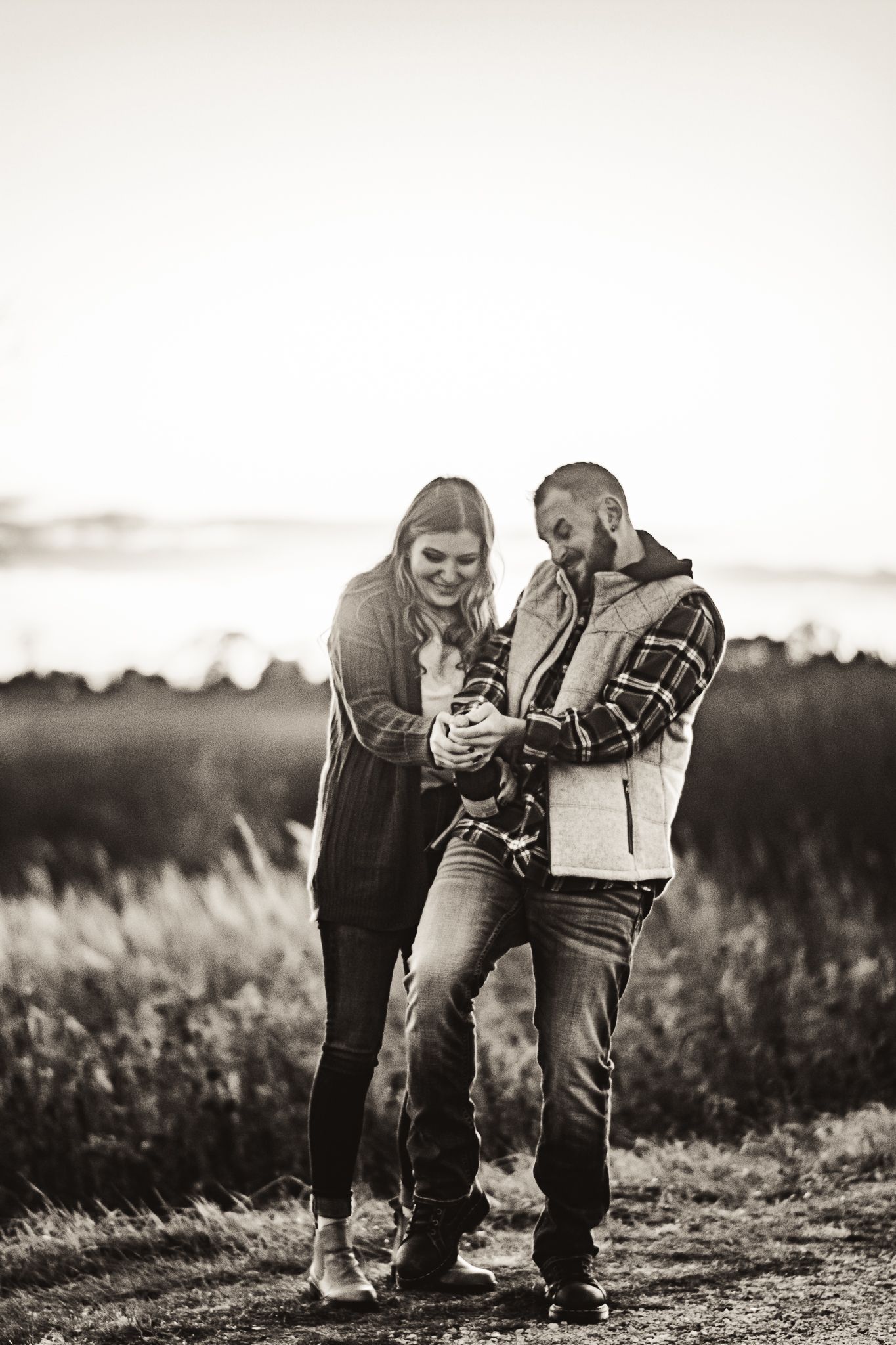 A happy couple shares a lighthearted moment while looking at something together in a scenic field at sunset.