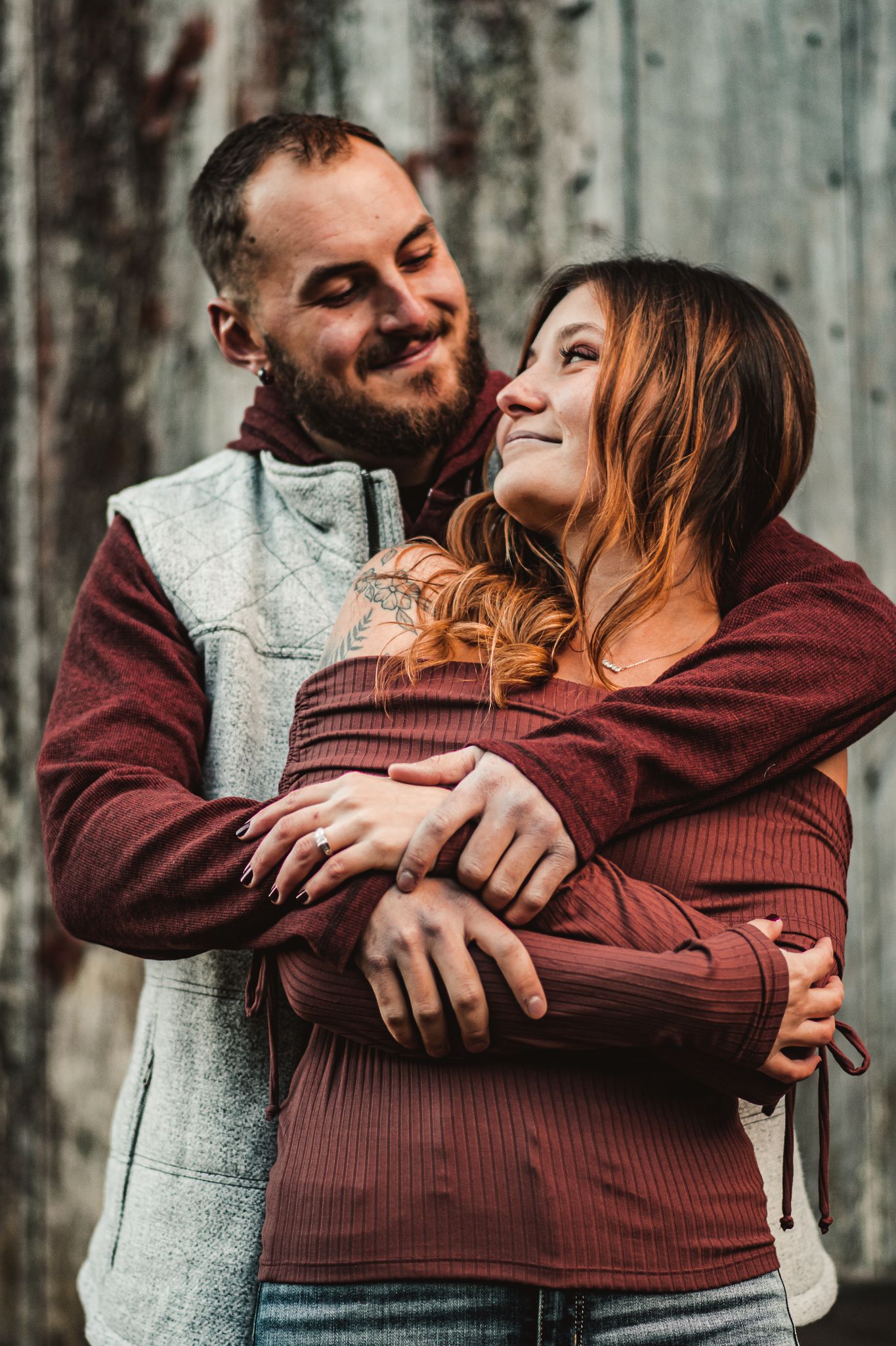 Intimate outdoor portrait of couple embracing in casual fall attire against wooden backdrop.