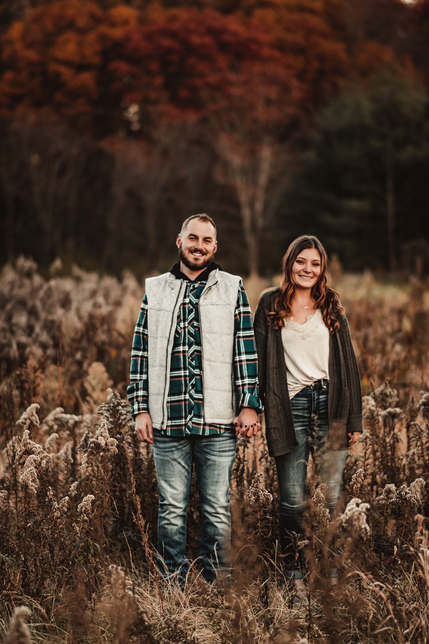 A couple stands together in a fall field wearing casual layered outfits and jeans.