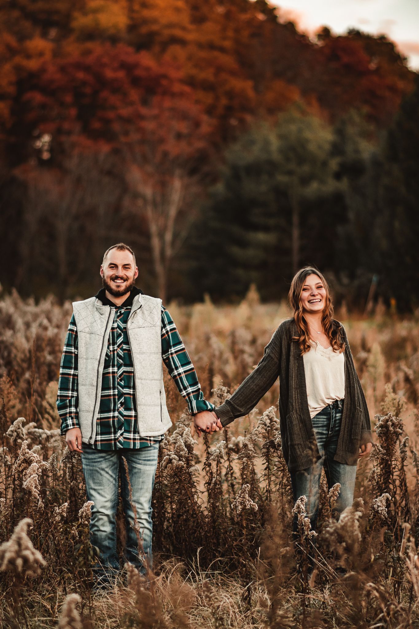 A couple holds hands while walking through a field of tall grass at sunset.