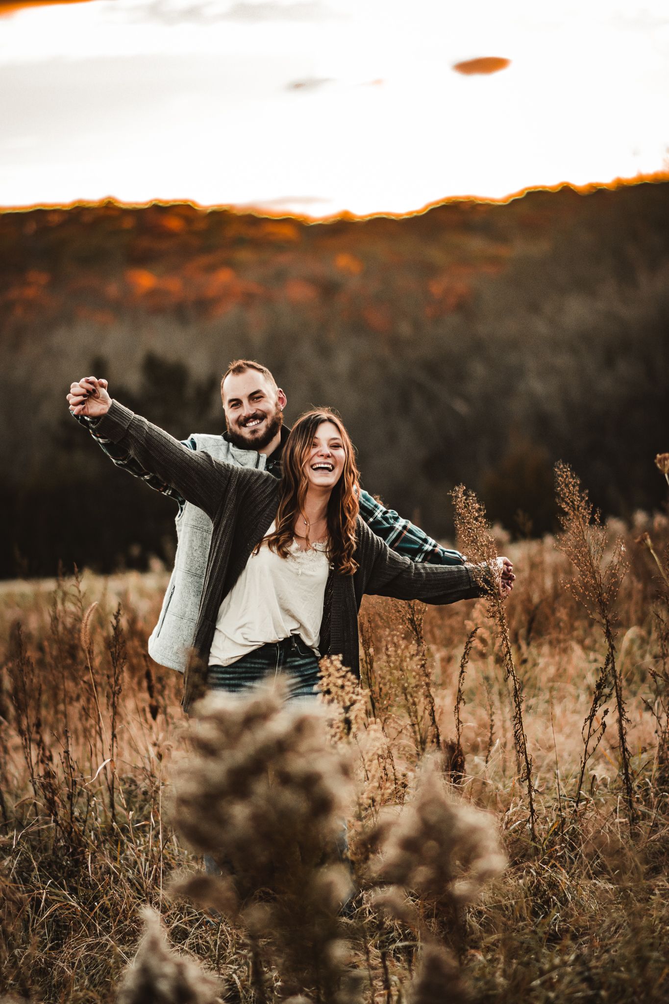 A couple spreads their arms with joy in a field during golden hour.