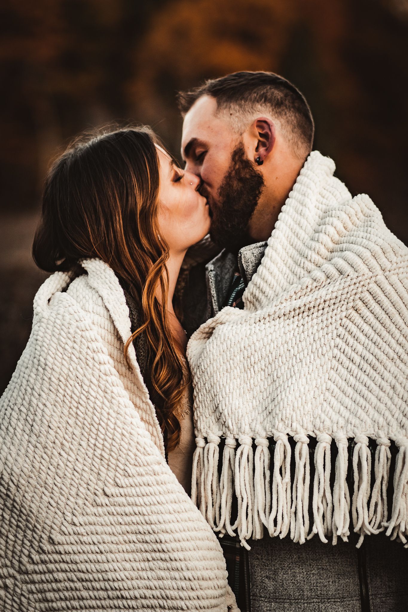Intimate couple sharing sweet kiss while wrapped in cream knit blanket during fall photoshoot.