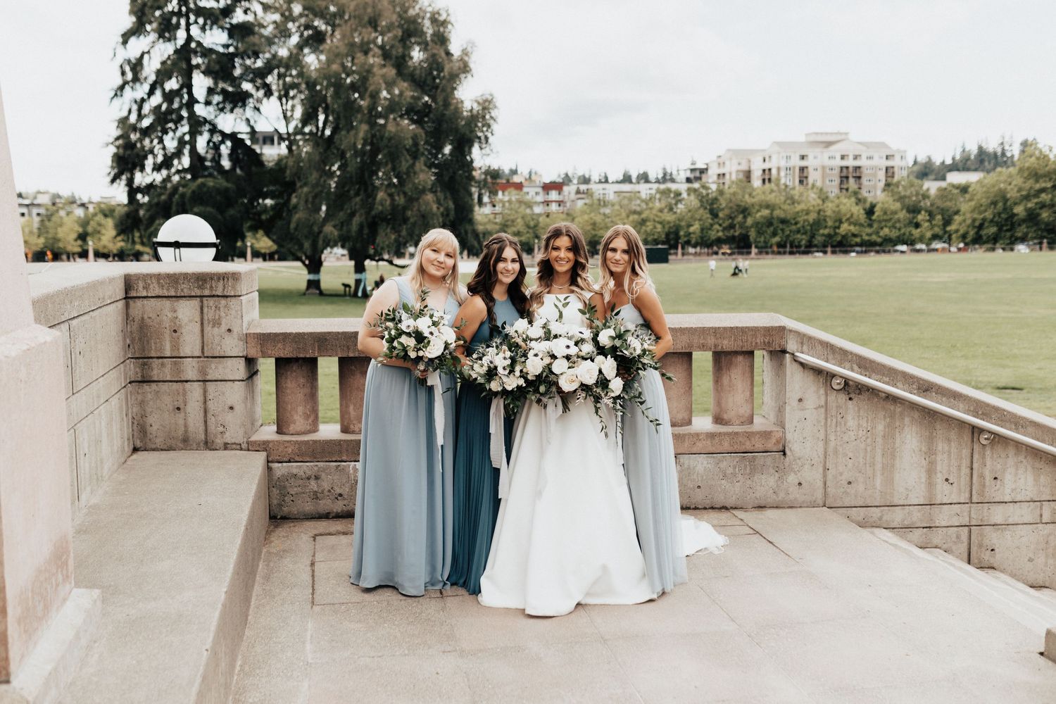 Bridesmaids in blue dresses pose with bride holding floral bouquets on stone terrace.