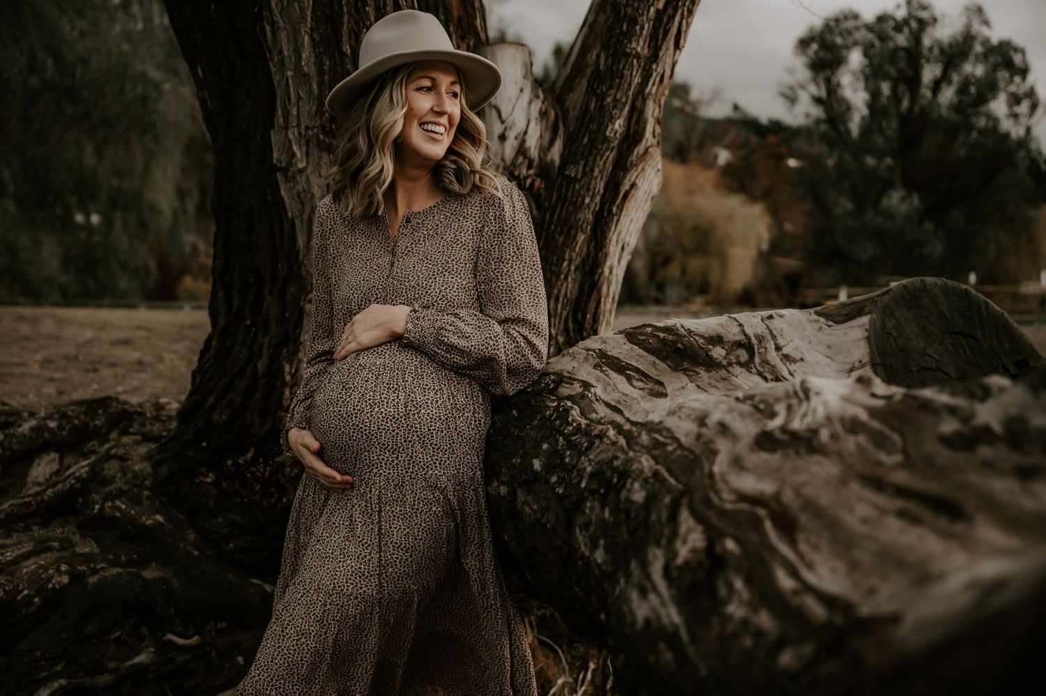 An expectant mother in a brown sweater dress and hat poses against a tree trunk in a moody outdoor setting.