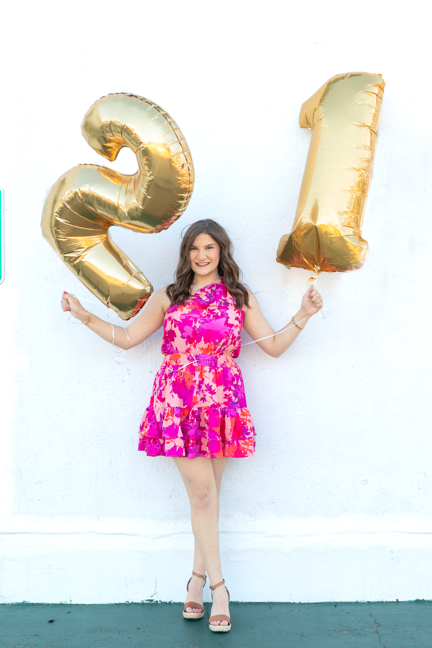 Young woman in pink floral dress holds gold number 21 balloons against white wall.
