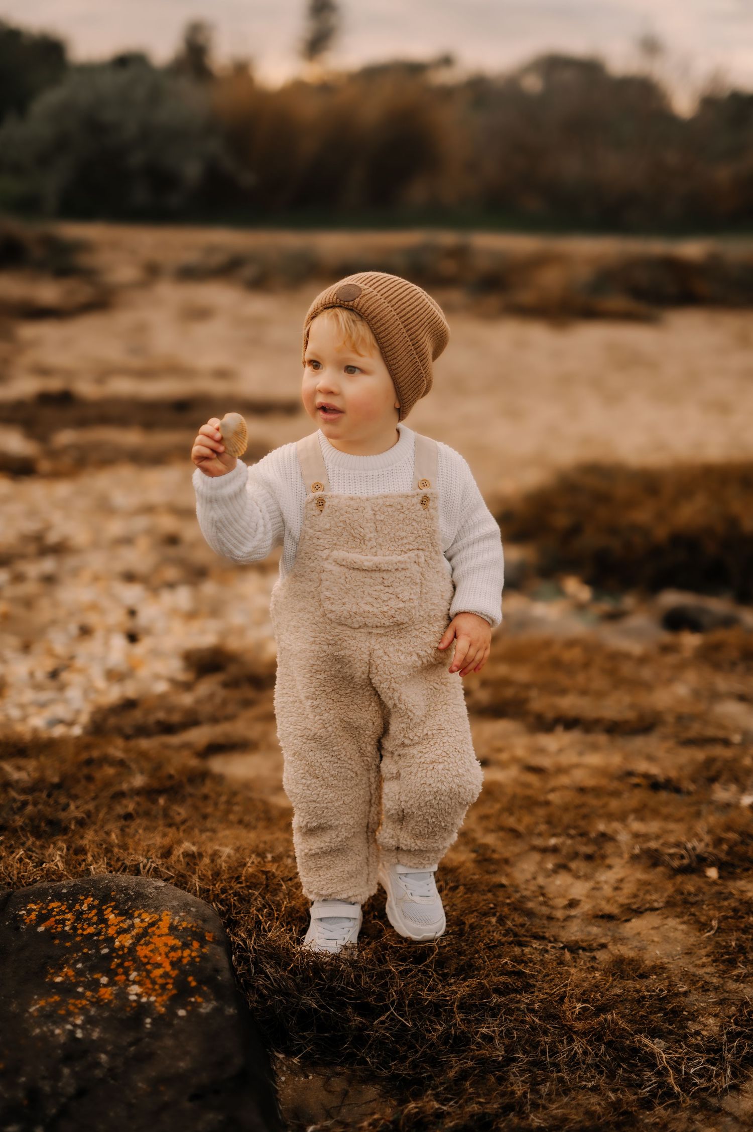 A toddler in beige overalls and brown beanie stands on rocky terrain during golden hour.