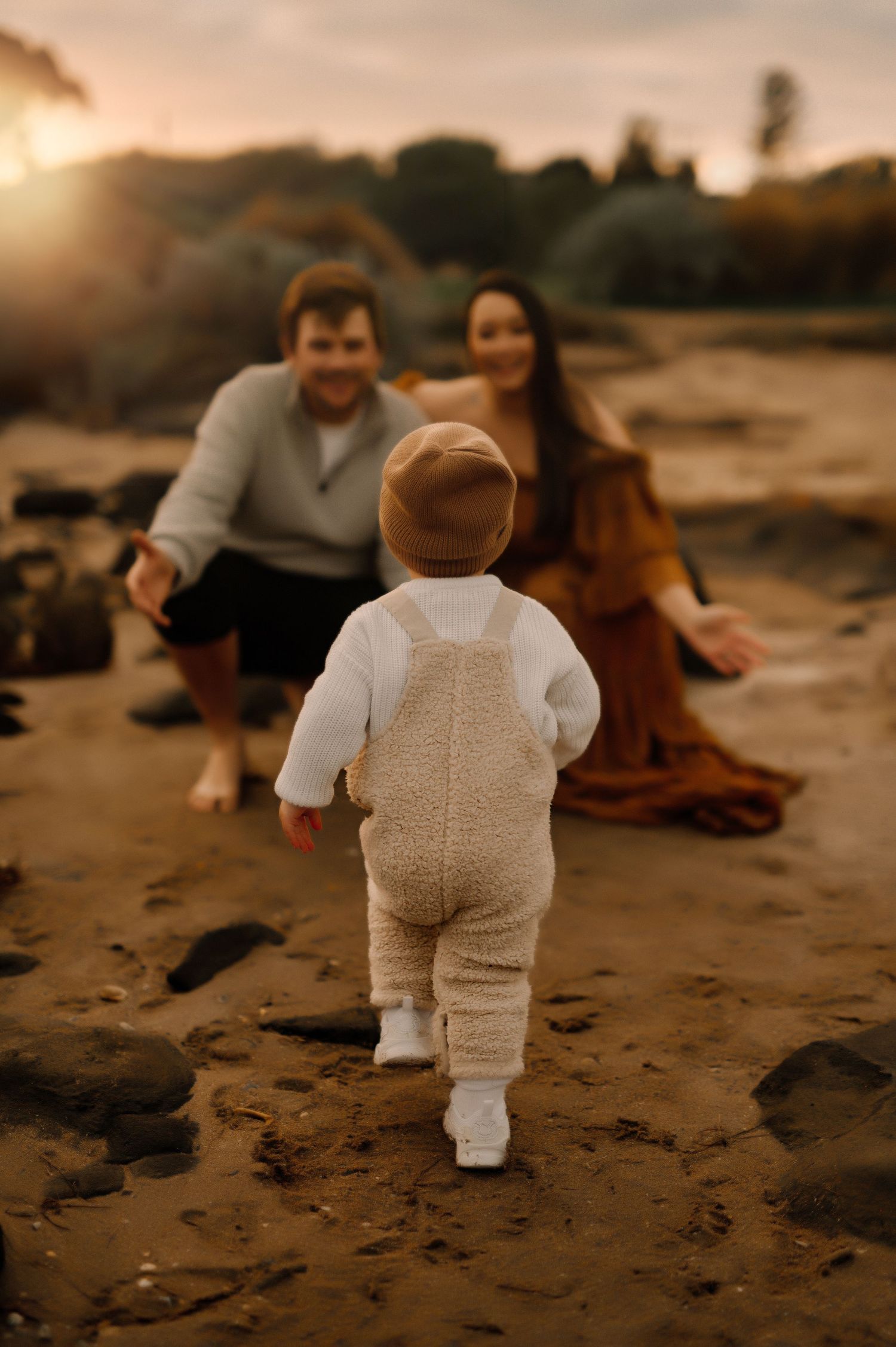 A toddler in white clothing walks towards adults sitting on rocky beach at sunset.