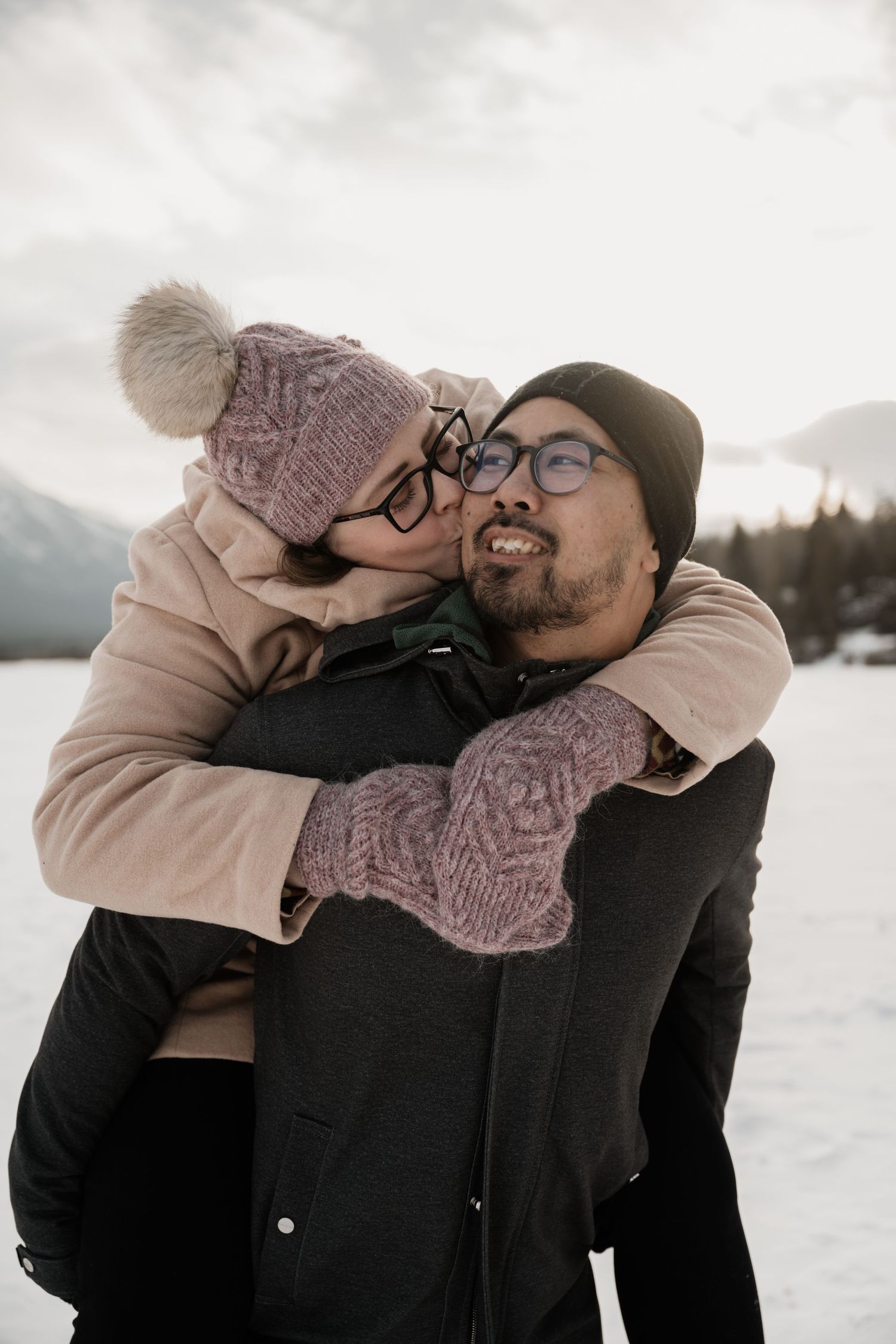A couple embraces in winter snow, wearing warm knitted hats and winter clothing against a mountainous backdrop.