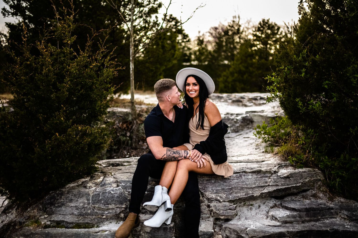A couple shares an intimate moment while sitting on rocky terrain surrounded by evergreen trees.