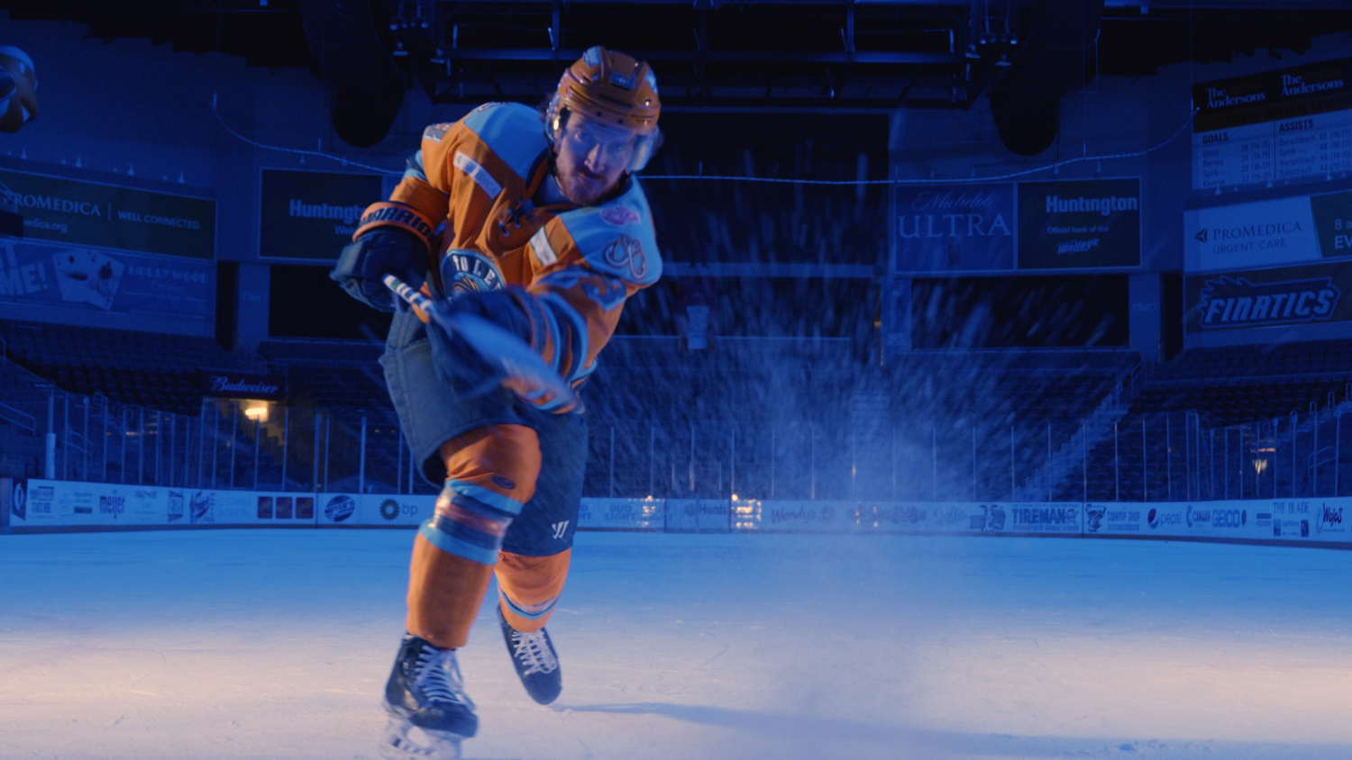 A hockey player in an orange jersey practices on illuminated ice.