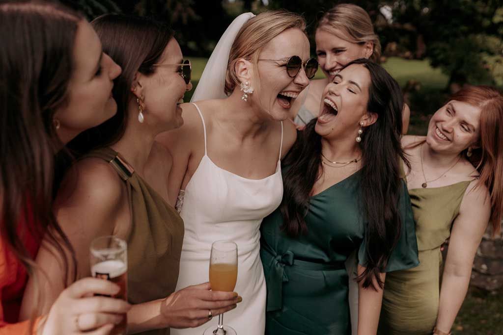 Group of friends in colorful dresses laughing and celebrating with drinks at an outdoor gathering.
