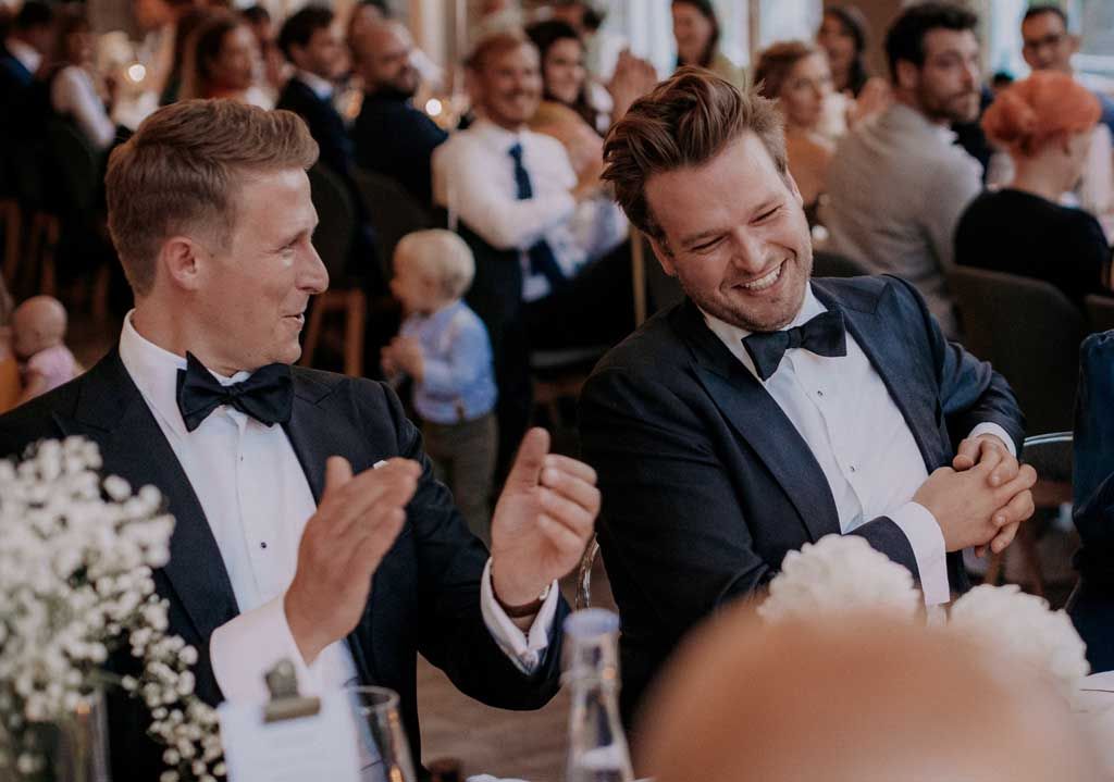Two groomsmen in tuxedos sharing a laugh during wedding festivities.