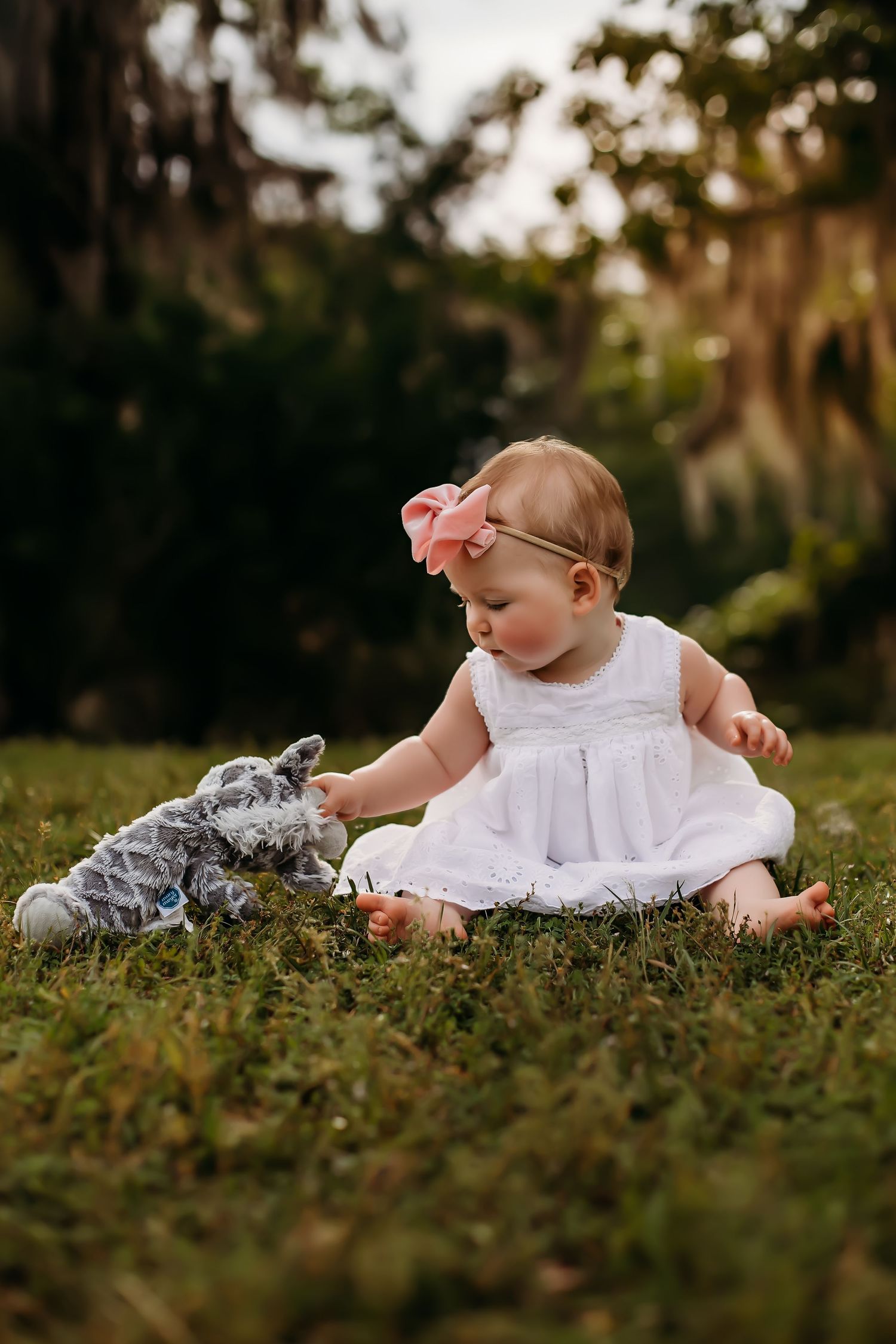 Baby in white dress playing with gray stuffed toy on grassy lawn.