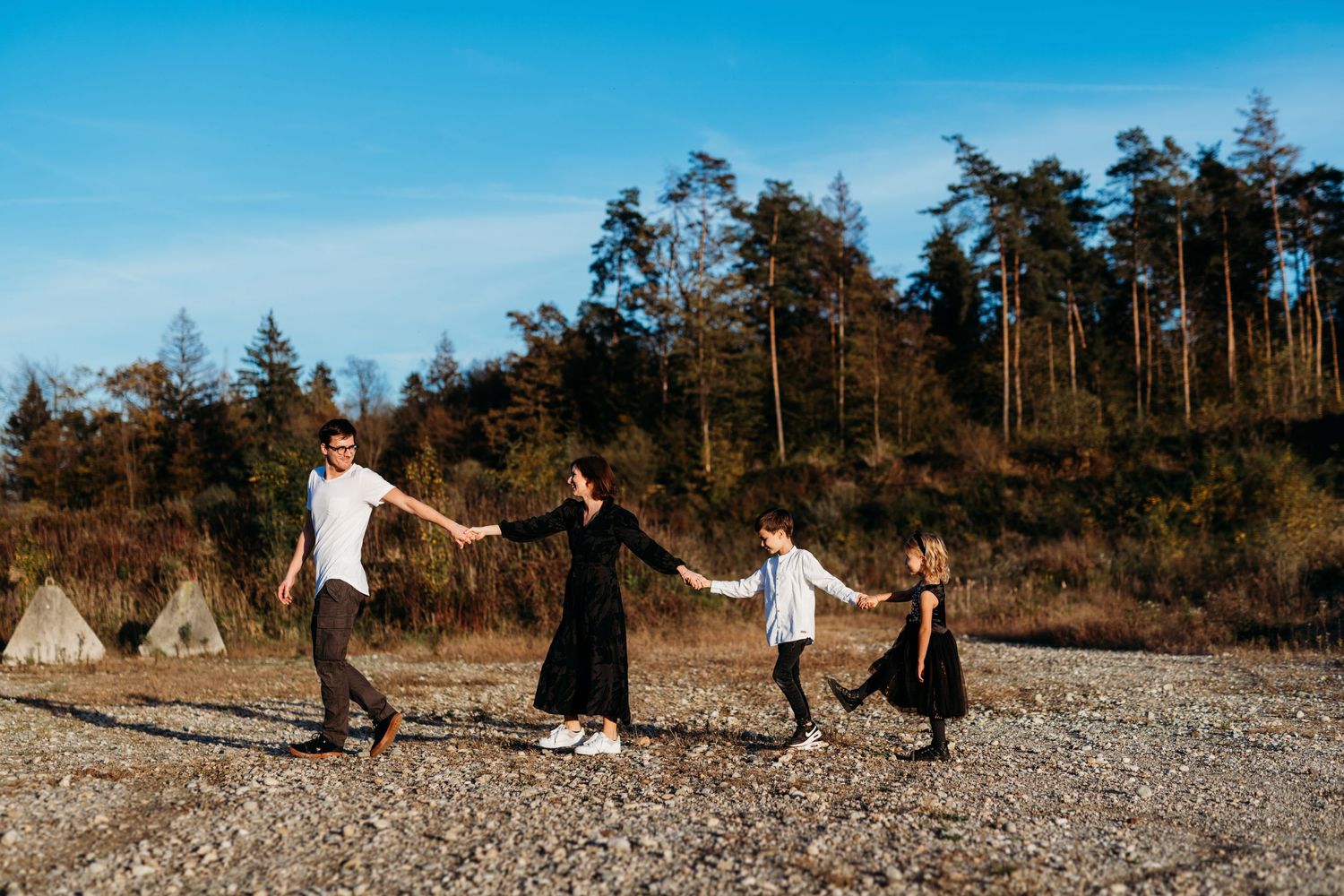 Four people hold hands and dance joyfully on a rocky beach with towering evergreens in the background.