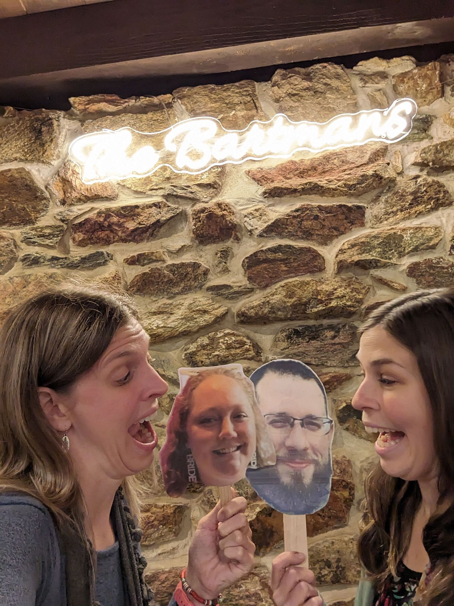Three friends pose playfully with a face cutout against a stone wall with illuminated signage.