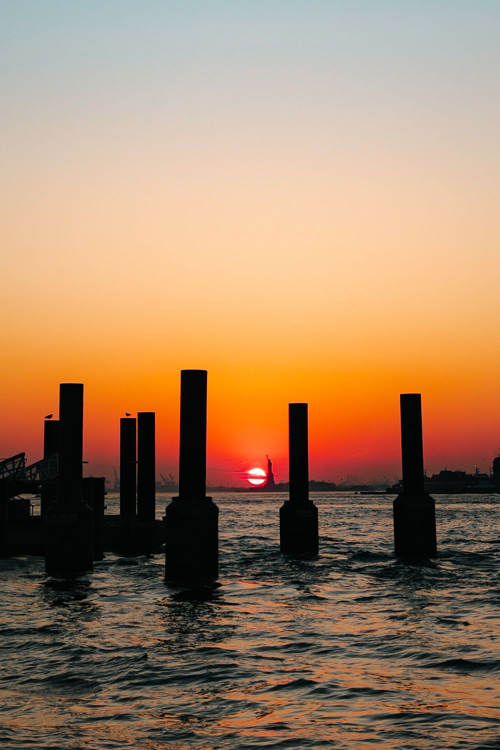 Silhouetted wooden posts stand in calm water against a vibrant orange and pink sunset sky.